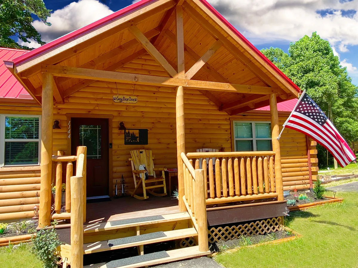 A warm exterior view of the log cabin, highlighting the inviting porch with comfortable seating and an American flag. The image suggests a welcoming entry and a perfect spot for gathering with family, enjoying coffee, or simply relaxing after a day of adventure.