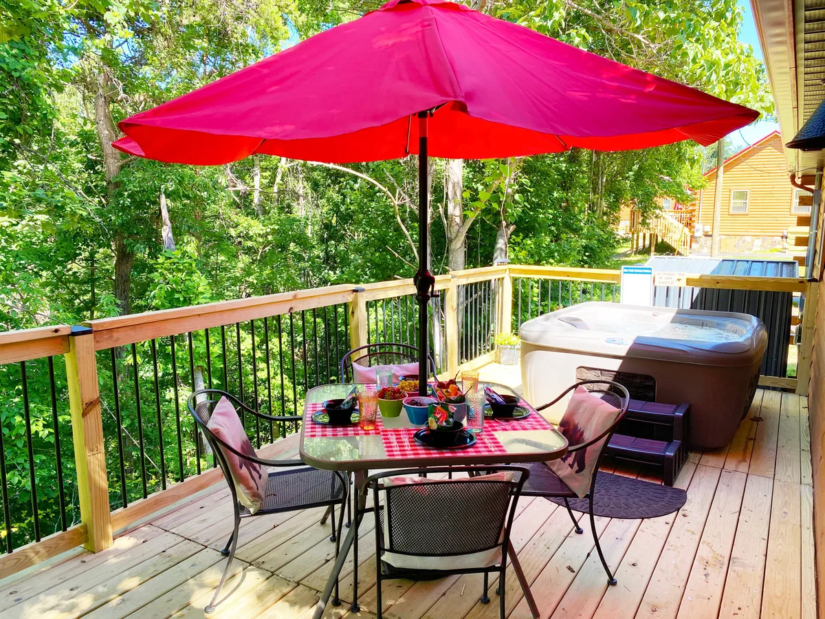 A cheerful outdoor setting featuring a large table with a red-checkered tablecloth, a bright red umbrella, and lush greenery all around. The hot tub in the background adds another fun amenity, while the setup illustrates opportunities for family meals and relaxation outdoors.