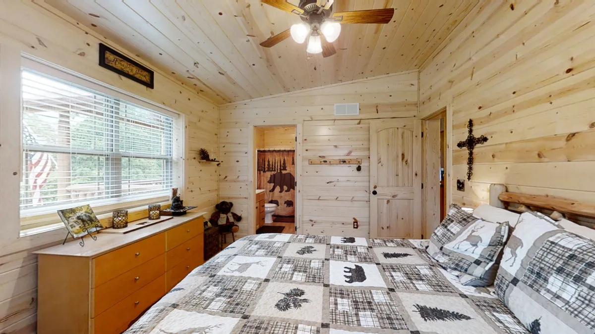 A restful bedroom with a bed dressed in a patterned quilt, wooden dresser, window with blinds, and rustic wood ceiling and walls. The room glows with natural light, offering a tranquil escape.