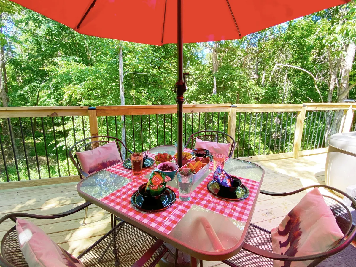 This image depicts an outdoor dining area with a red umbrella, checkered tablecloth, and plates set for a cheerful meal. The green foliage in the background enhances the inviting, festive mood, perfect for alfresco dining or a romantic brunch.
