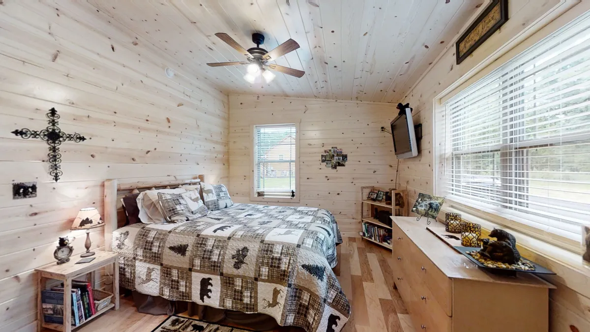 A cozy bedroom featuring a bed with a patterned quilt, wooden furniture, a decorative cross, and a TV mounted on the wall. Warm natural light fills the space, creating a welcoming and relaxing atmosphere for guests.