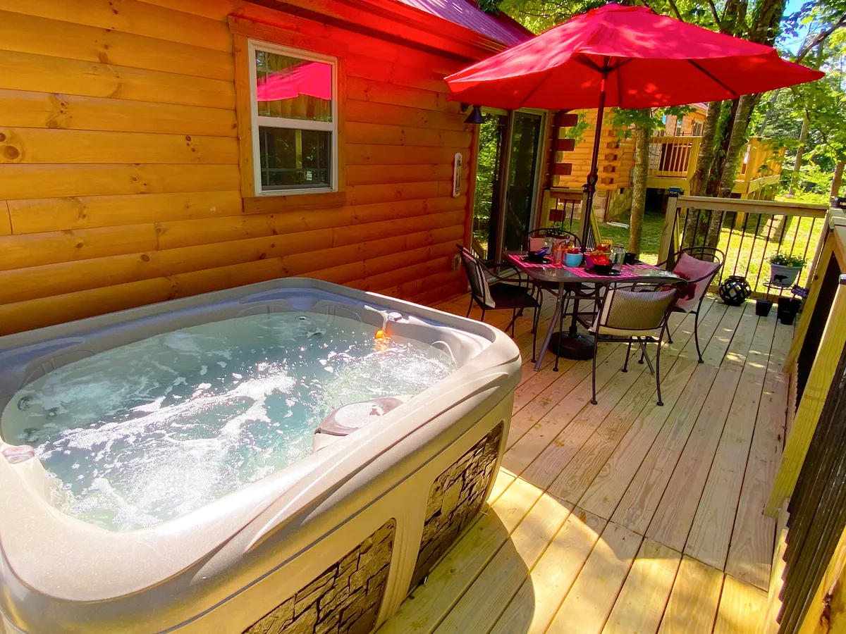 This image showcases the back deck, featuring a bubbling hot tub and an inviting outdoor dining table shaded by red umbrellas. The space is surrounded by greenery, creating a peaceful, private atmosphere perfect for relaxation and socializing.