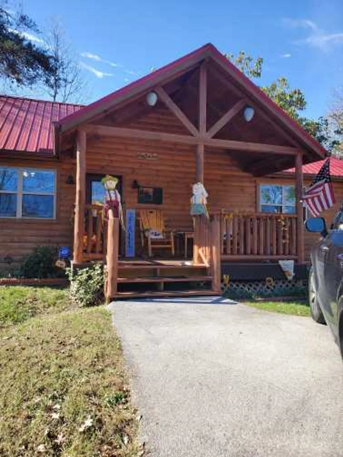 This image captures the welcoming front porch of Cozy Bear Den, featuring comfortable rocking chairs, wooden details, and homey seasonal decorations. The porch looks out toward leafy greenery, offering a glimpse of the tranquil, private setting. The American flag and scarecrow décor add to the inviting, charming atmosphere.