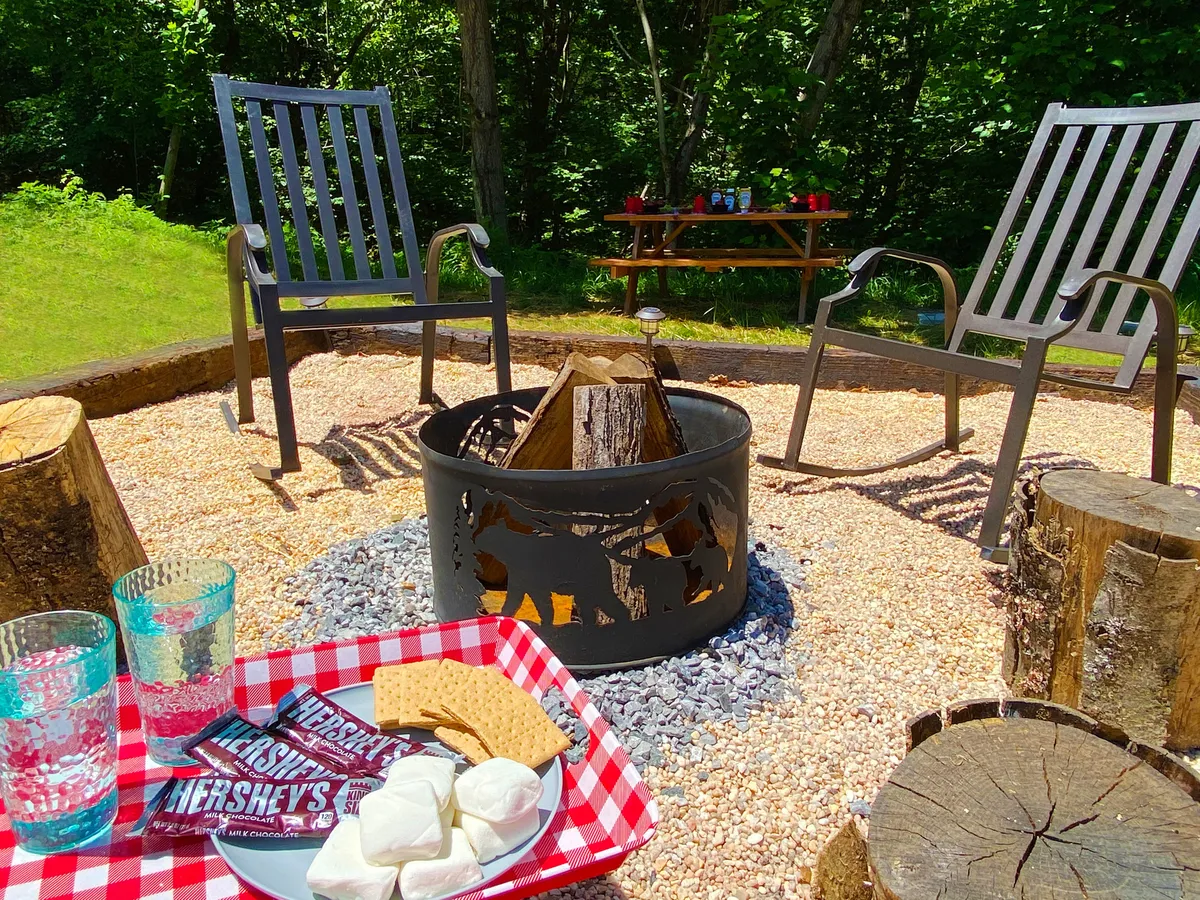 A cozy fire pit area with camp chairs and wooden log seating is set up for making s'mores. S'mores ingredients are visible, and the scene suggests a relaxed evening ambiance ideal for gathering and storytelling under the stars.