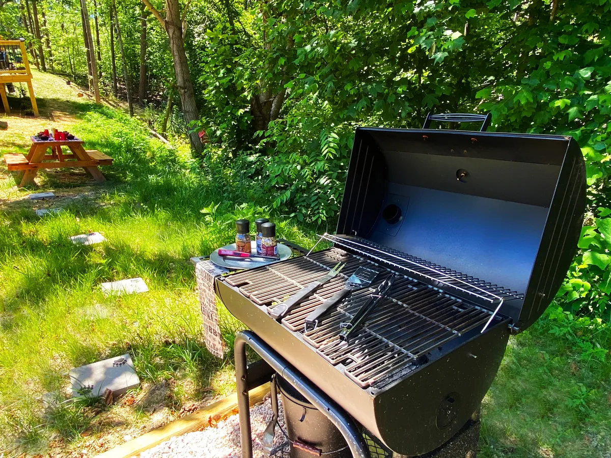 A relaxed outdoor setup featuring a grill, seasoning jars, and a wooden picnic table, inviting scenes of barbecuing and shared meals. The sunlight and simple decor enhance the sense of casual group fun and togetherness.