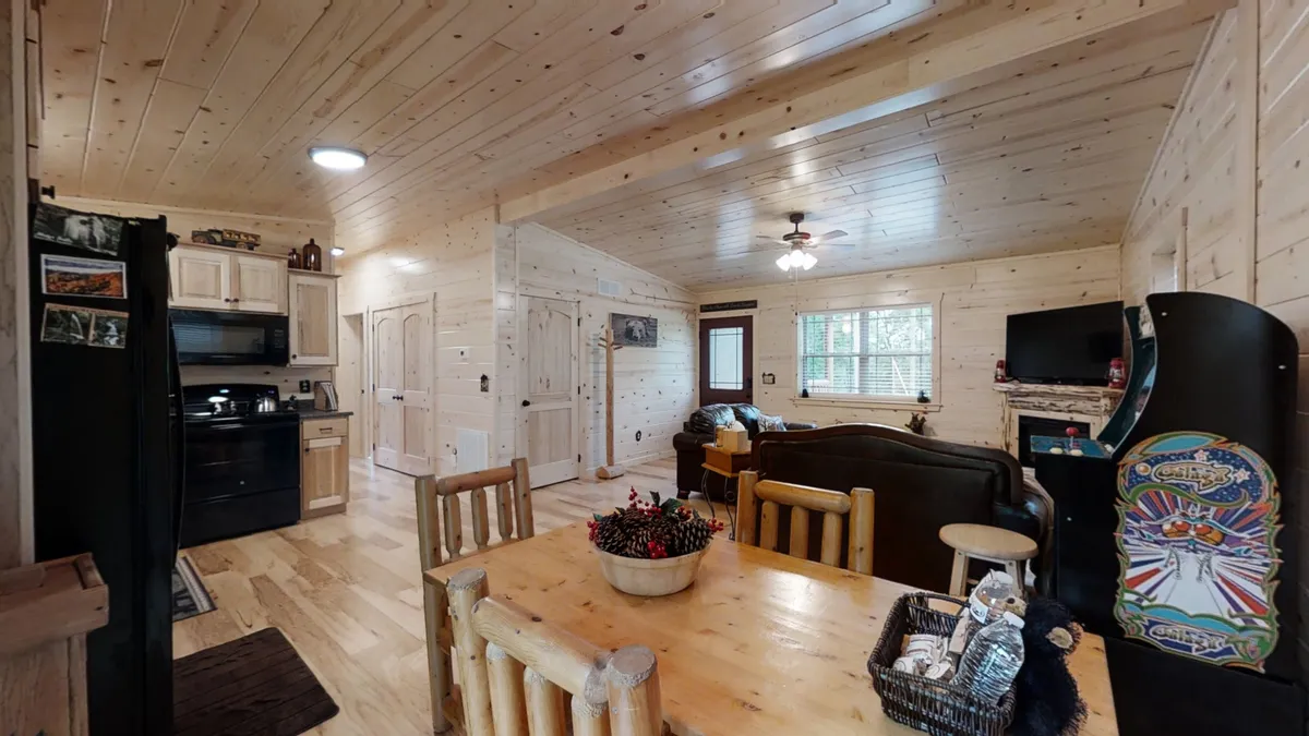 This image provides a slightly different angle of the dining and living areas, featuring a dining table, sofa, kitchen appliances, and an arcade game machine. The setting exudes warmth and is designed for both group interaction and relaxation.