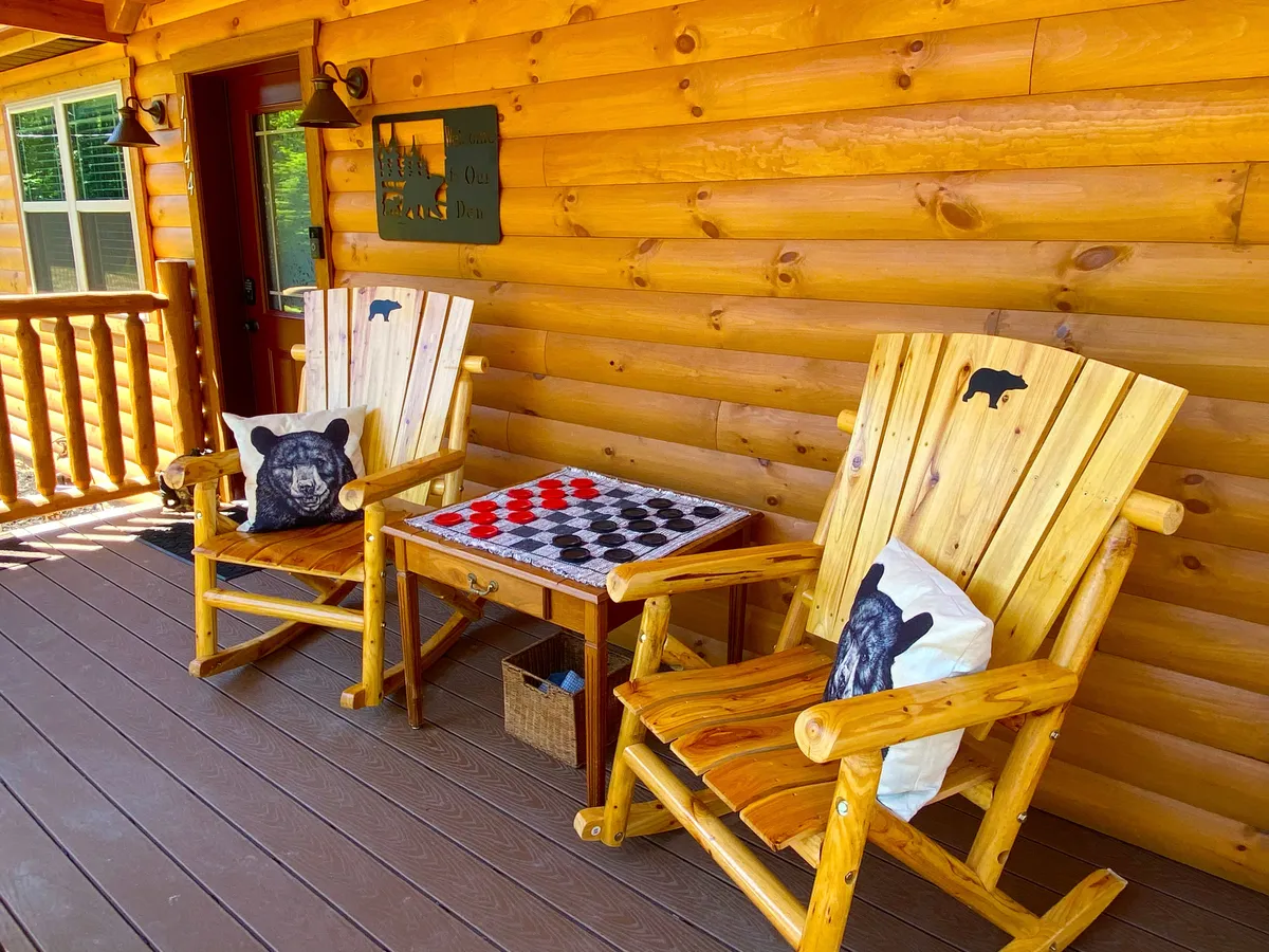 A cozy exterior shot of the log cabin’s front porch, featuring classic wooden rocking chairs, a checkers game table, and bear-themed pillows. The porch looks out onto peaceful greenery, inviting guests to savor coffee or cocktails while enjoying mountain views.