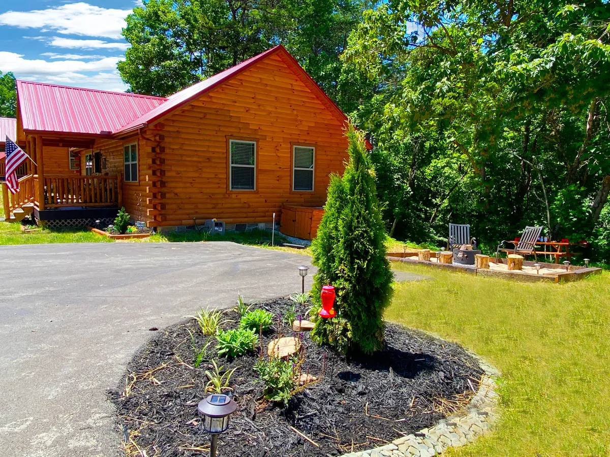This exterior shot captures the charming log cabin with a flower bed in front, a visible flag, and a welcoming fire pit area surrounded by seating. The yard offers plenty of space for outdoor family games and gatherings, with a cozy, inviting atmosphere that promises memorable evenings around the fire.