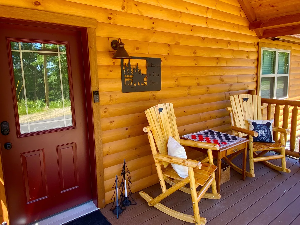 The front porch features classic rocking chairs, a checkerboard table, and a charming red door, all set against the backdrop of mountain air and natural light. The scene feels cozy and invites guests to relax and enjoy the view or play games.