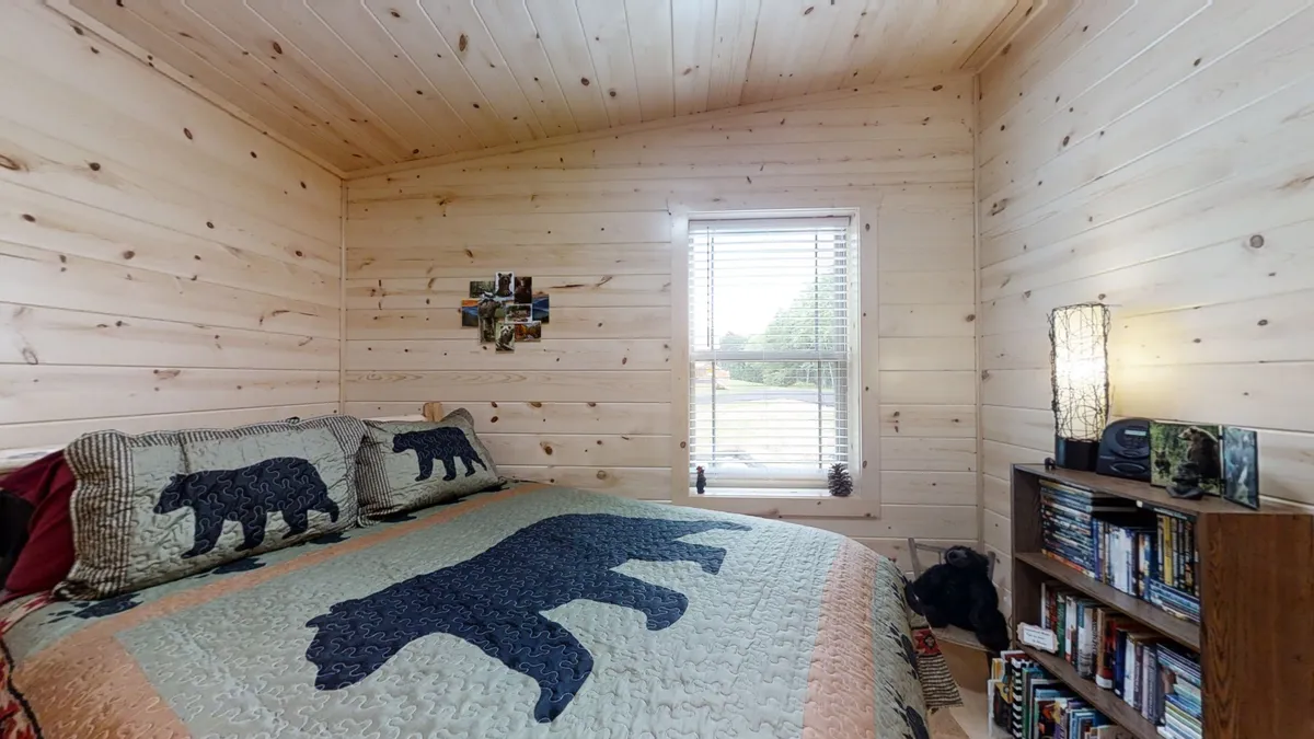 A charming bedroom with a bear-themed quilt, rustic wood paneling, a window letting in natural light, and a cozy bookshelf. The mood is warm and inviting, perfect for children or friends.